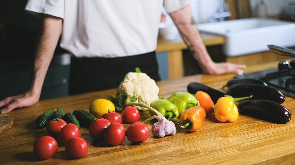 Diverse fresh vegetables on kitchen countertop with a person in the background. Perfect for healthy lifestyle content.